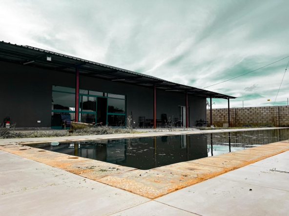 A modern building with large glass windows under a cloudy sky, fronted by a still water reflecting its facade, with a concrete walkway leading to the entrance.